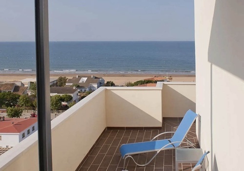 Balcony sea view room at Hotel Pato Amarillo, a 4-star hotel in Punta Umbría, Huelva. Blue lounge chair overlooking beach & ocean.