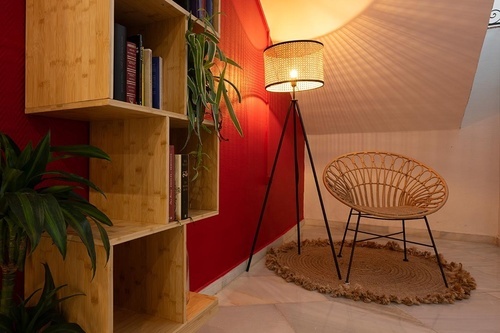 Relaxing corner at Pato Rojo Hotel in Punta Umbria with a rattan chair, lamp, and wooden bookshelf.