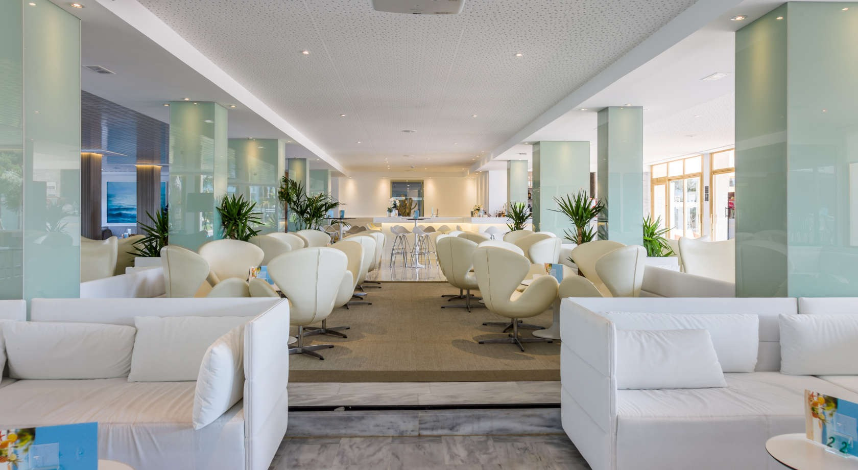 Salón luminoso del hotel con sofás blancos y sillas huevo, barra de bar al fondo, ideal para Playa Esperanza Resort.