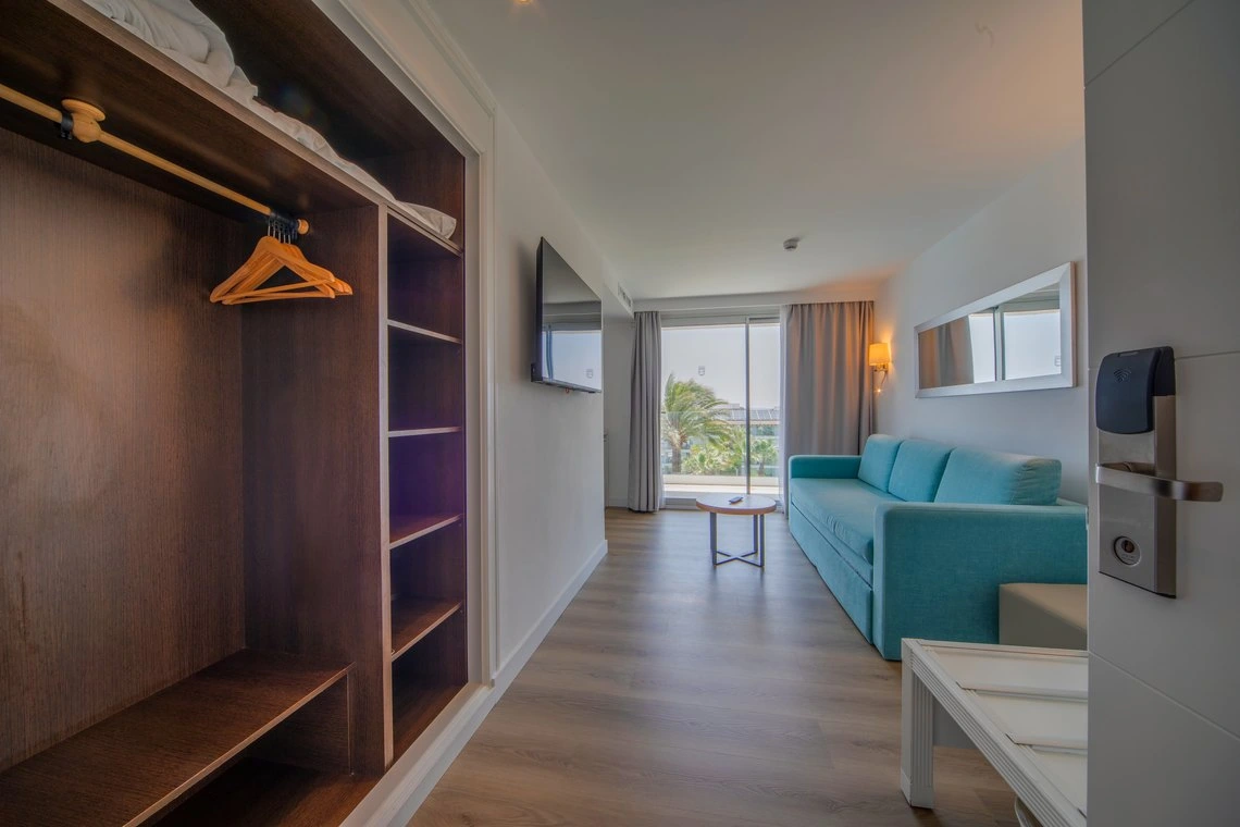 Amplia habitación de hotel con sofá azul claro, armario de madera y vista al exterior. Playa Esperanza Resort