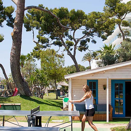 Jóvenes jugando al ping-pong al aire libre en el hotel con una cabaña y un hotel al fondo. Playa Esperanza Resort