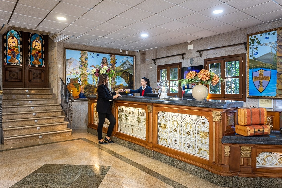 a woman stands at a hotel reception desk holding a drink