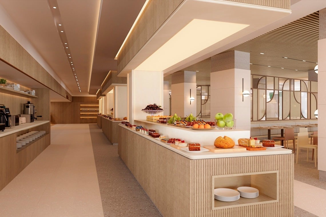 Un amplio y luminoso bufé moderno presenta una variedad de postres, frutas y panes frescos, con una estación de café a la izquierda y un comedor al fondo.