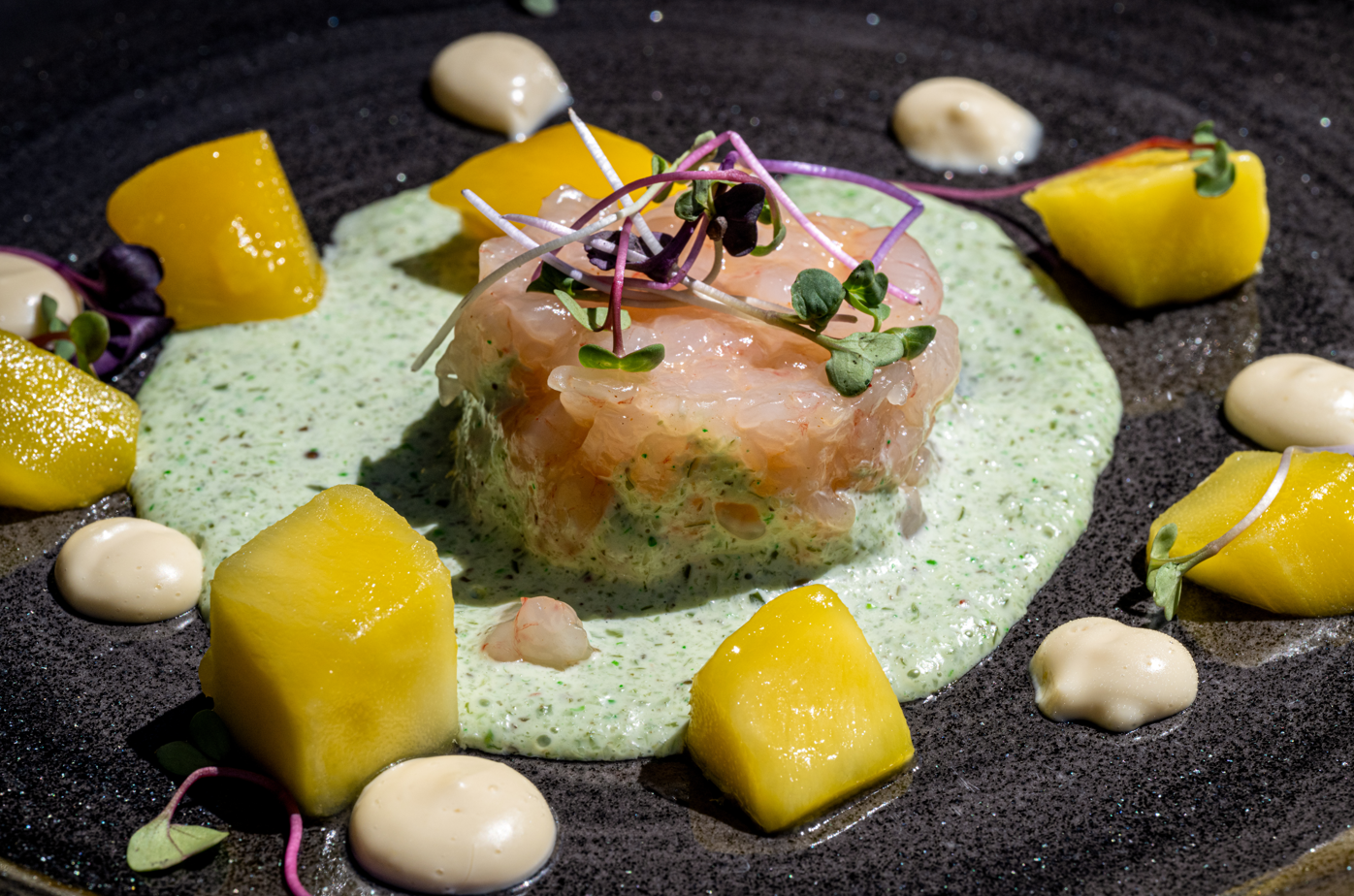 Elegant shrimp tartare with mango, a vibrant green sauce, and creamy garnishes on a dark plate, hotel dining.