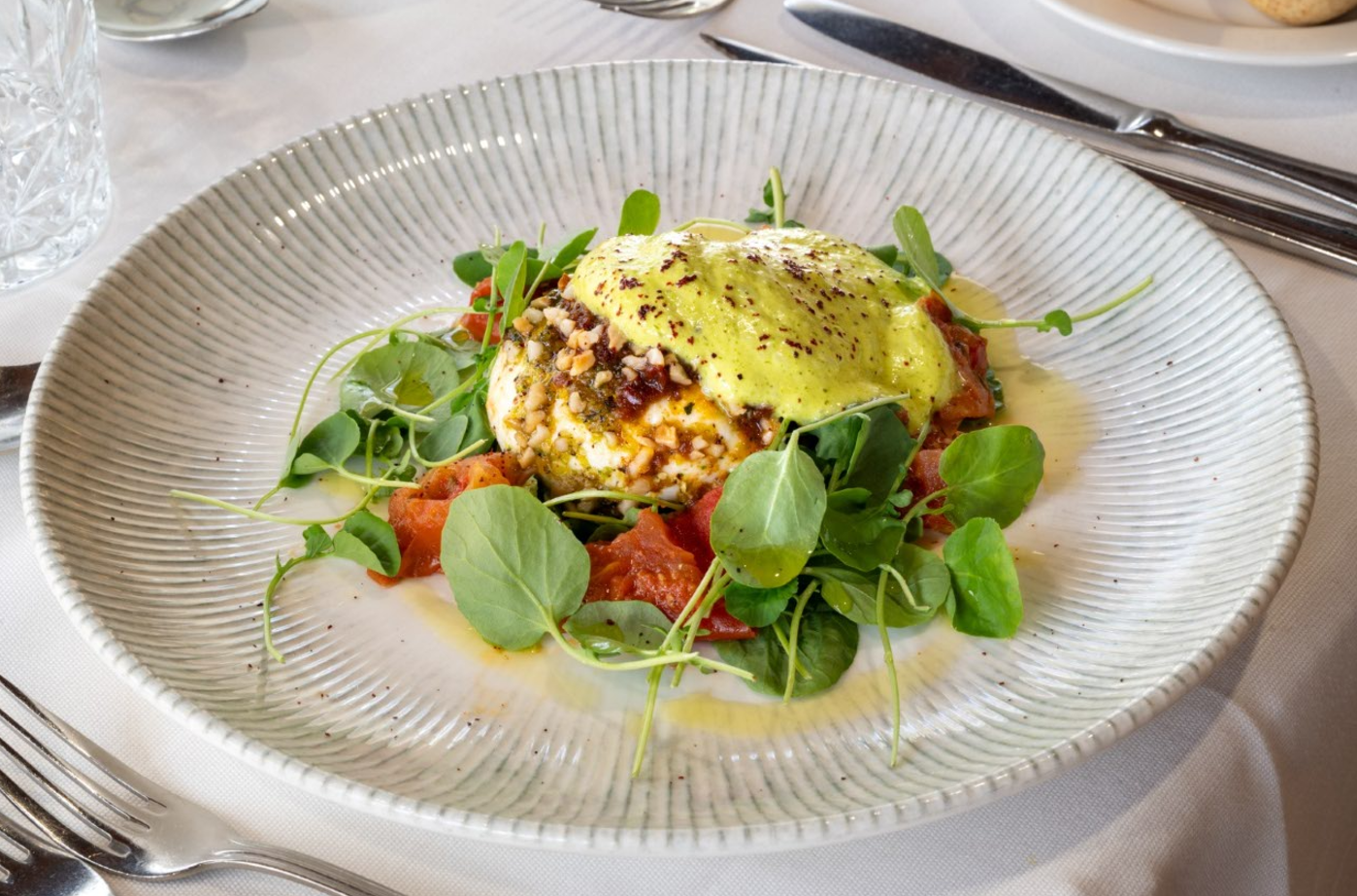 Un plato blanco con textura acanalada contiene un queso cremoso coronado con salsa verde, frutos secos y especias, rodeado de tomates rojos y hojas verdes.