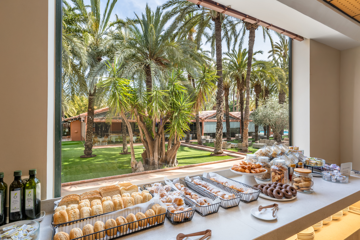 Un buffet de desayuno con pan y bollería se encuentra junto a un gran ventanal con vistas a un exuberante jardín lleno de palmeras y edificaciones al aire libre.