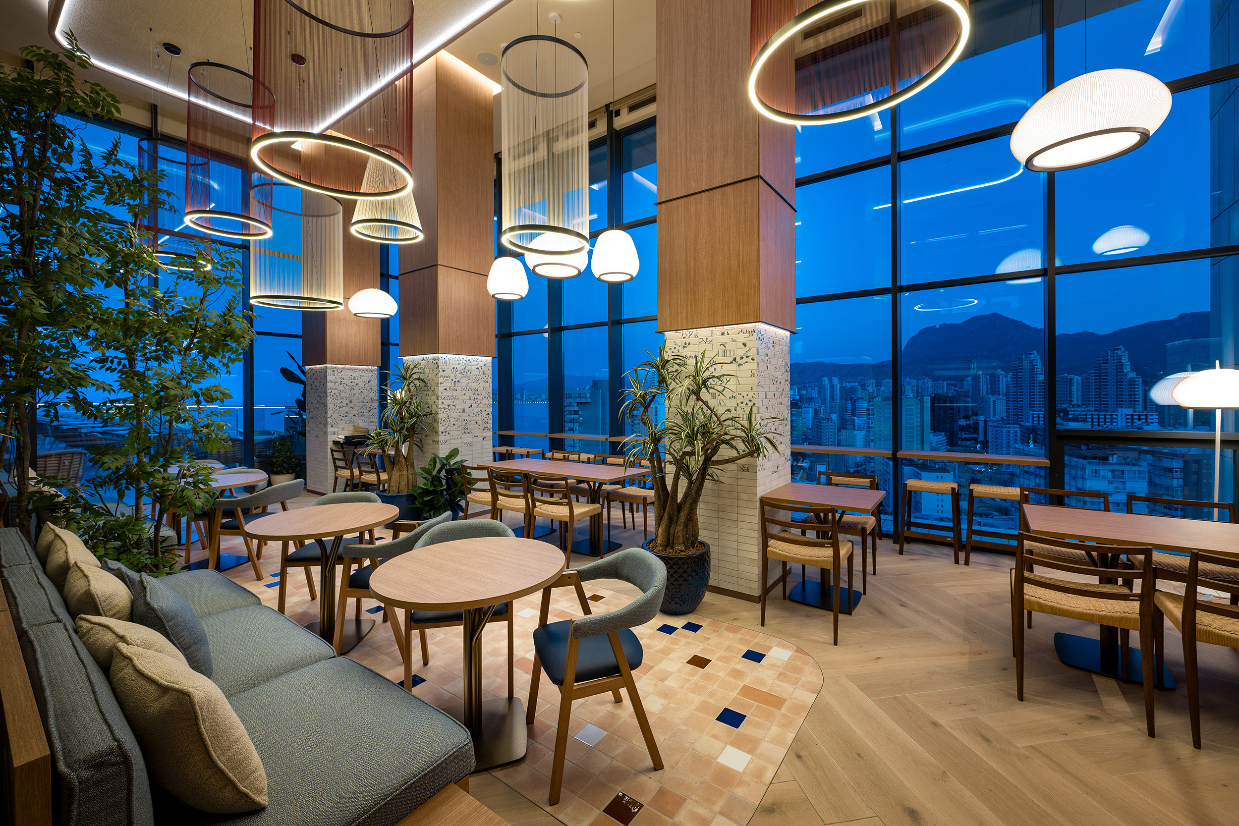Vista de restaurante moderno en hotel con luces colgantes, plantas y vista a la ciudad. Port Hotels: Corporativa