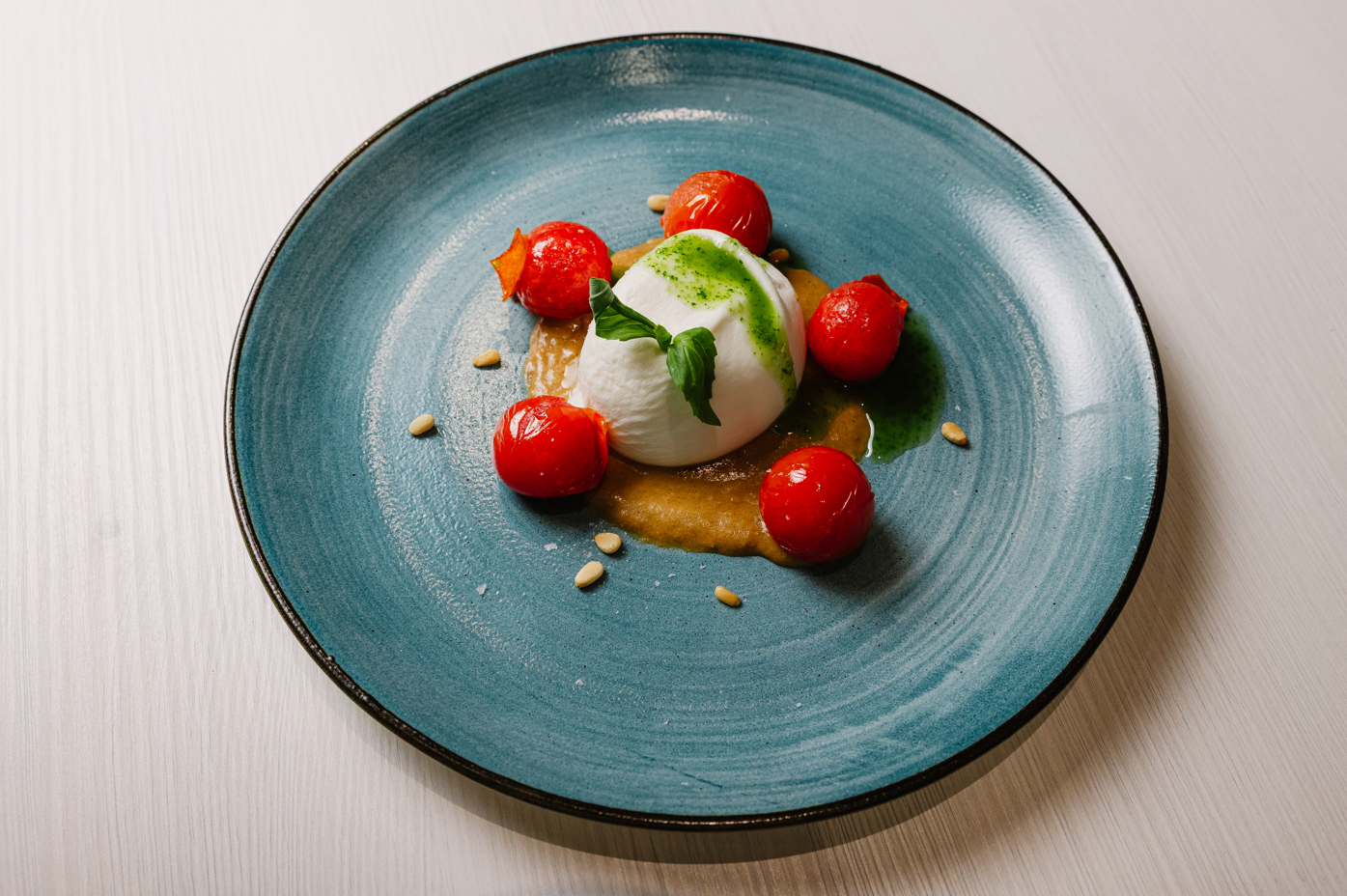 Exquisite burrata with basil pesto, roasted cherry tomatoes, and pine nuts on a vibrant blue plate.