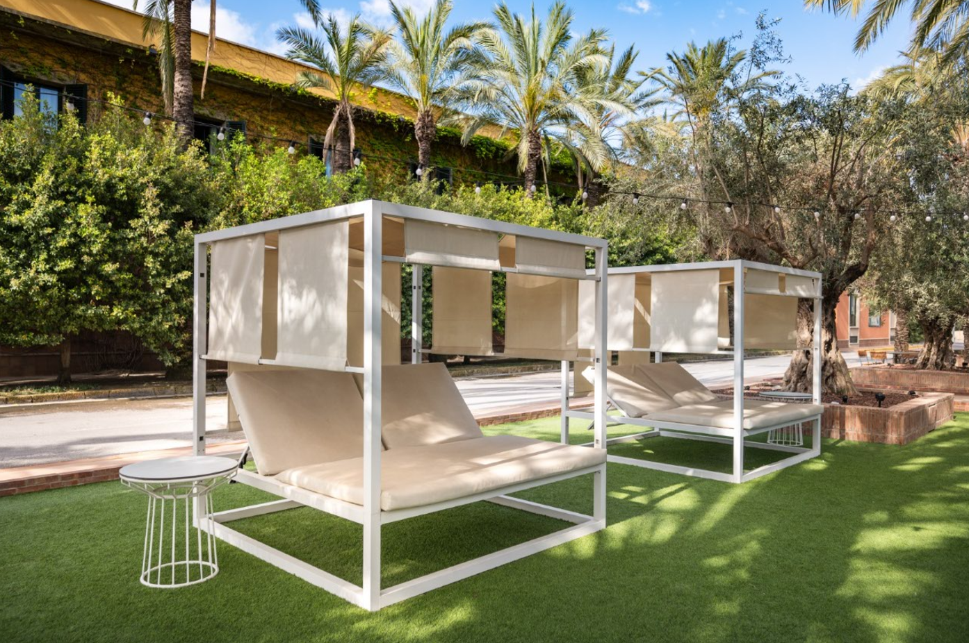 Dos tumbonas modernas con dosel y cojines beige se sitúan sobre césped artificial verde en un jardín tropical con palmeras y un edificio cubierto de hiedra al fondo.