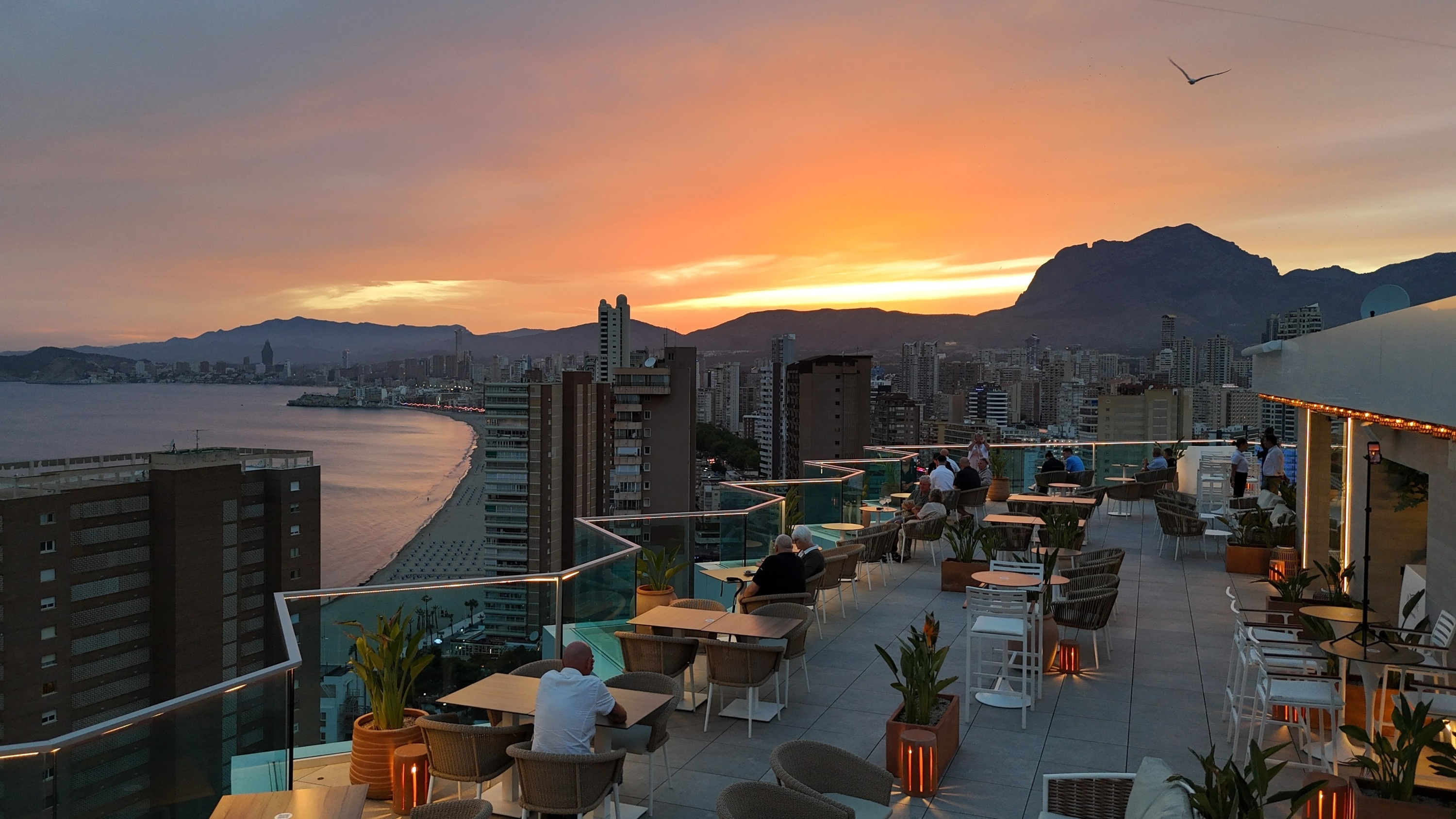 people sitting at tables on a balcony overlooking a city at sunset