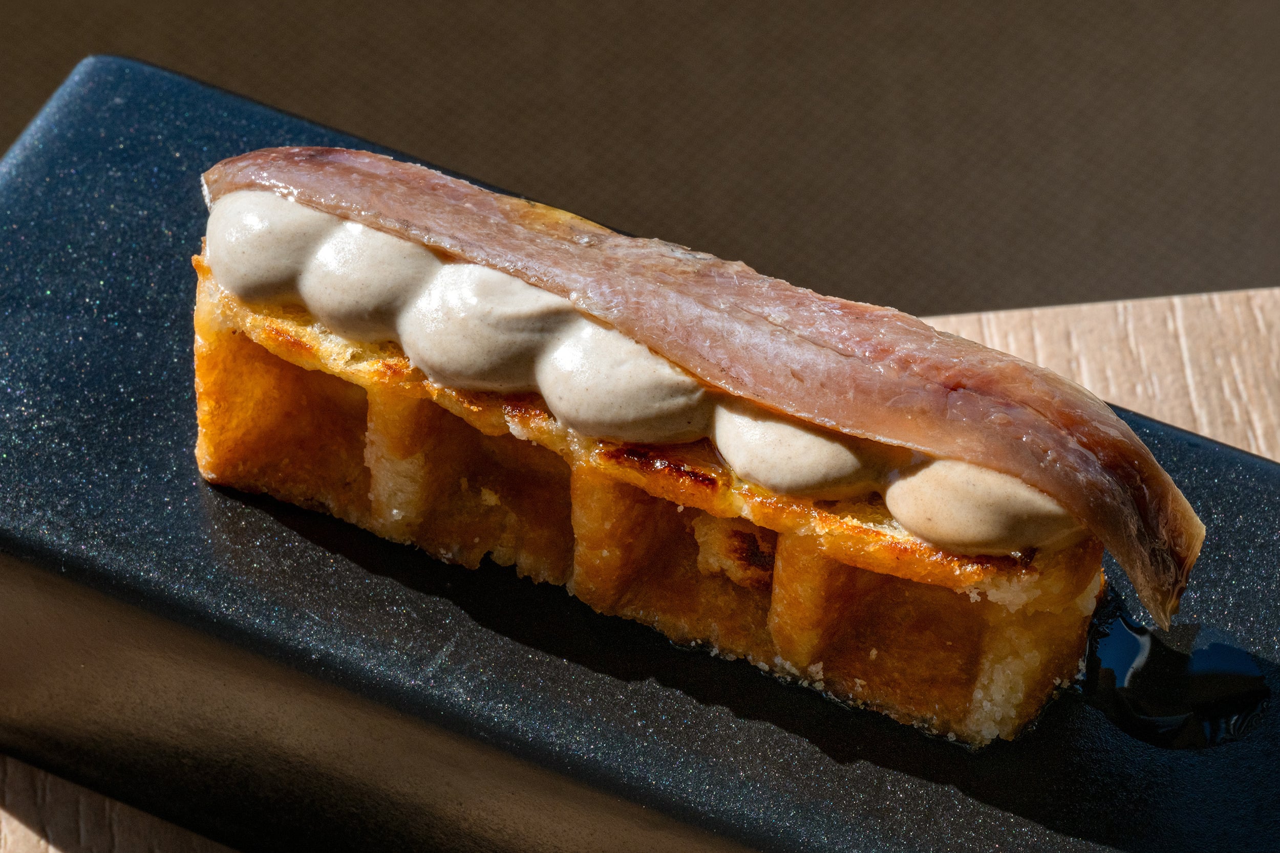 Port Hotels: Corporativa: tapa gourmet de anchoas y crema sobre pan frito en plato oscuro.