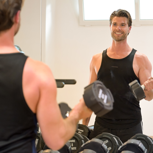 un hombre sostiene una mancuerna frente a un espejo en un gimnasio