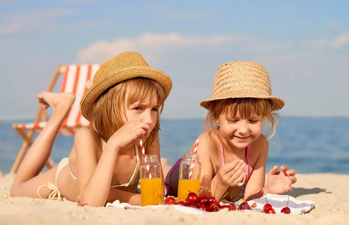 dos niñas acostadas en la playa comiendo cerezas y bebiendo jugo de naranja
