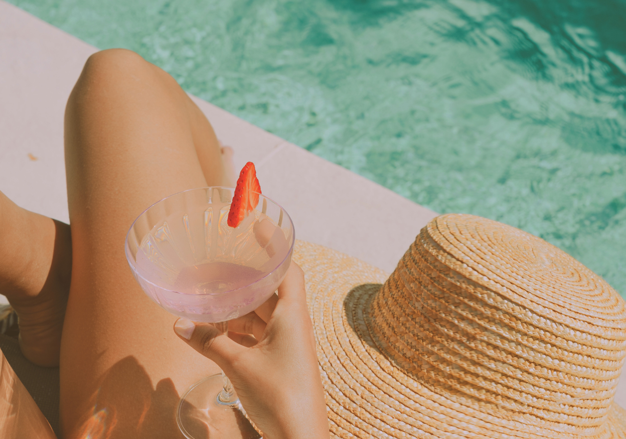 a woman in a straw hat sits by a pool holding a glass of pink liquid with a strawberry in it