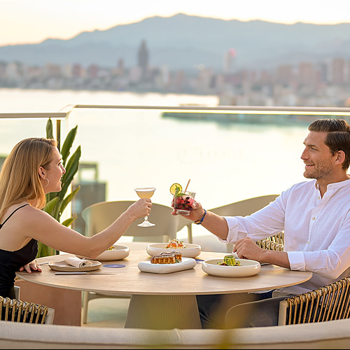 Pareja brindando con cócteles en una terraza con vistas al mar, ideal para Port Hotels: Corporativa.
