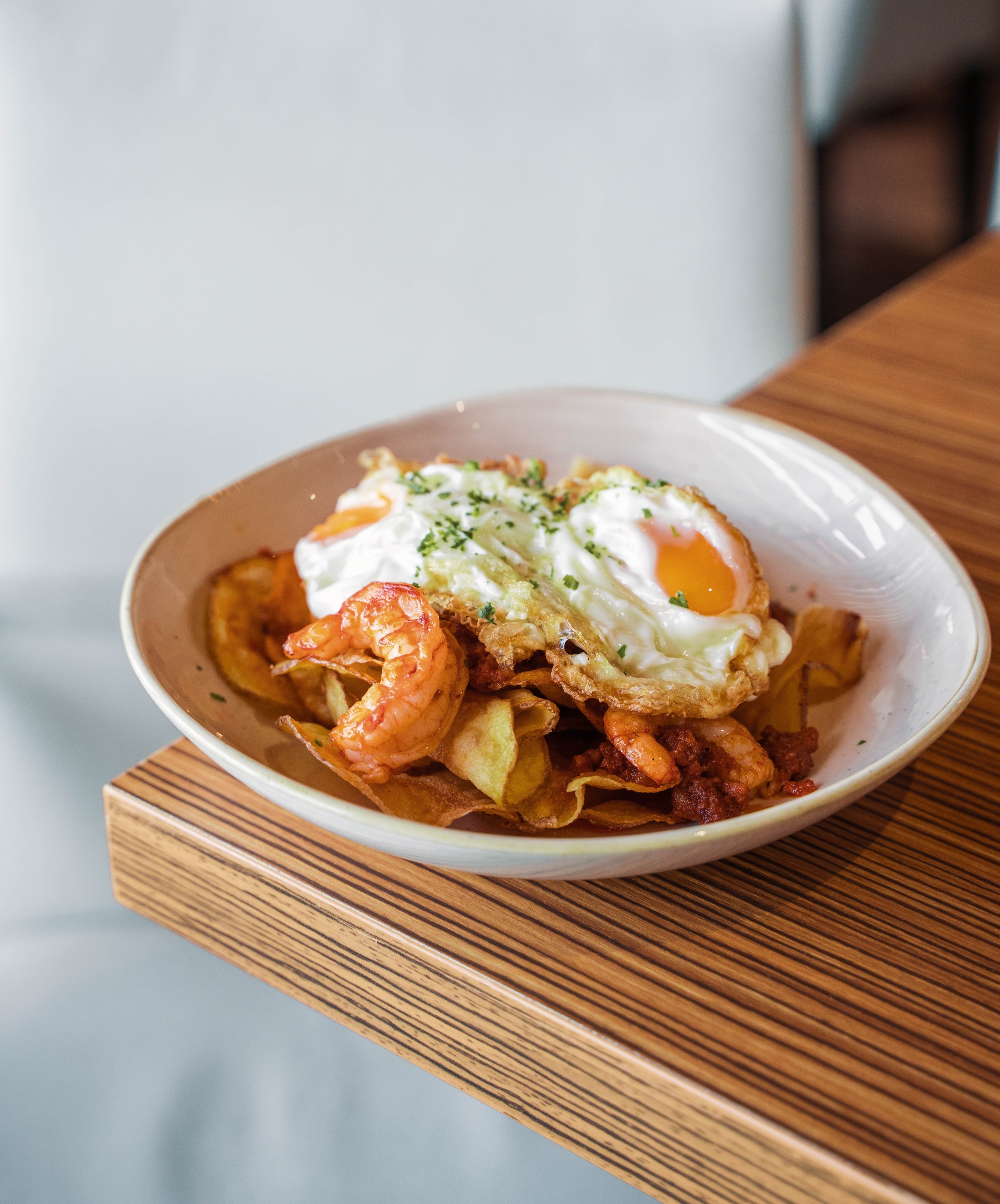 Hotel restaurant meal: fried eggs, shrimp, and crispy potatoes in a bowl on a stylish wooden table.