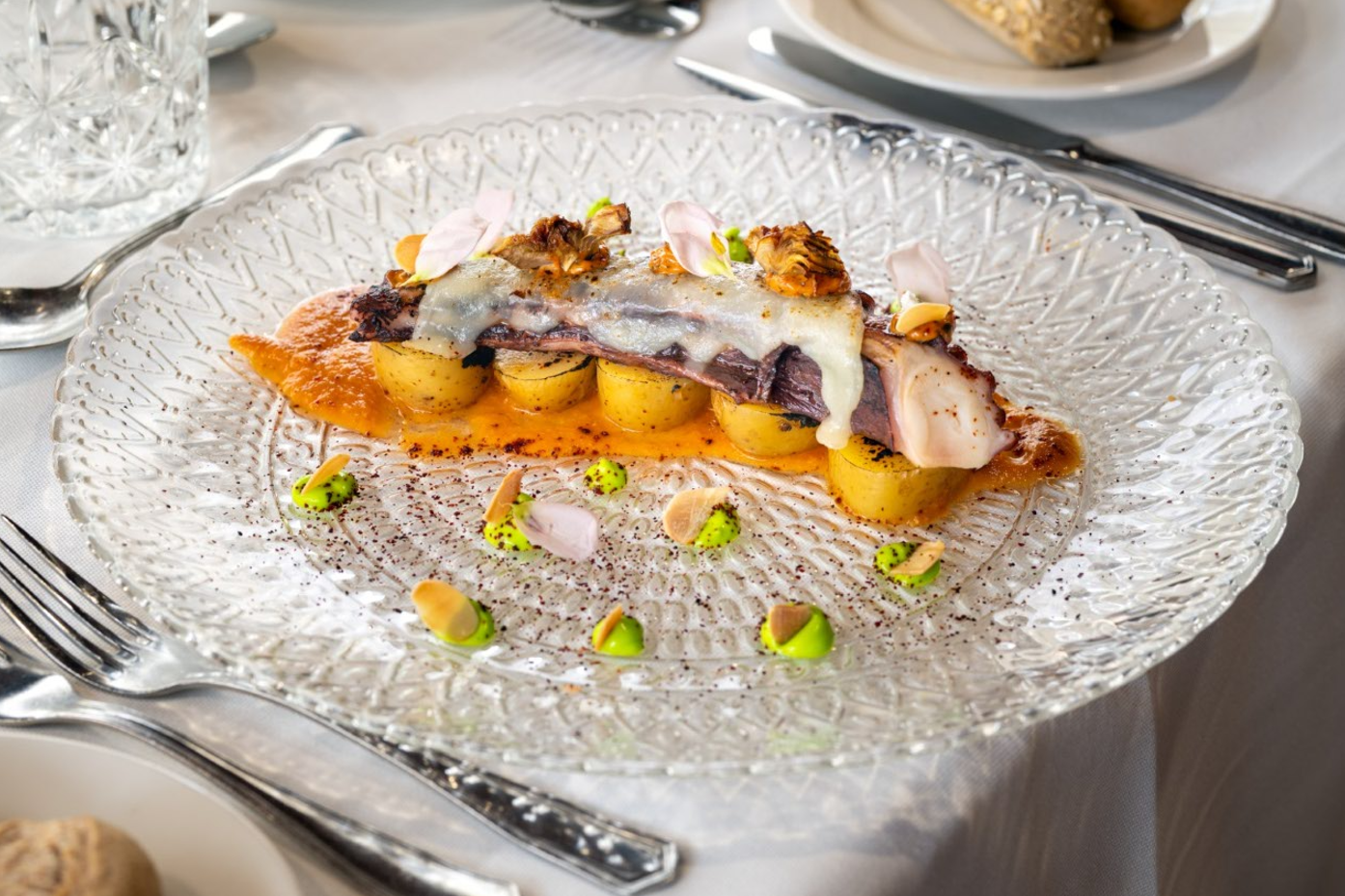 Un plato gourmet de pulpo con patatas, queso fundido, salsa anaranjada y adornos verdes y florales, presentado en un elegante plato de cristal.