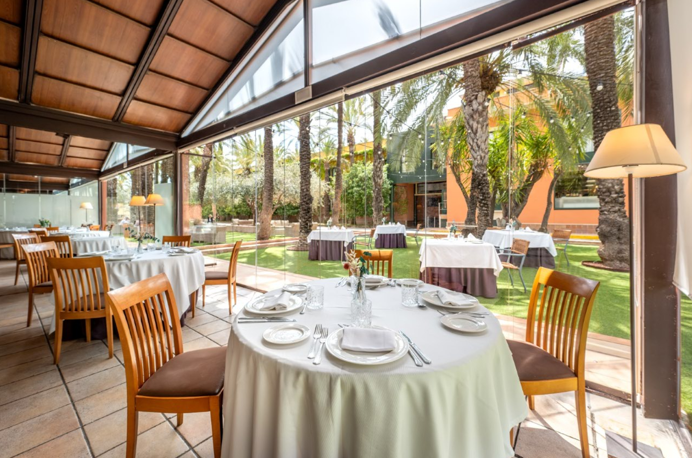 Un luminoso comedor de restaurante acristalado, con mesas puestas, ofrece vistas a un jardín tropical con palmeras y césped.