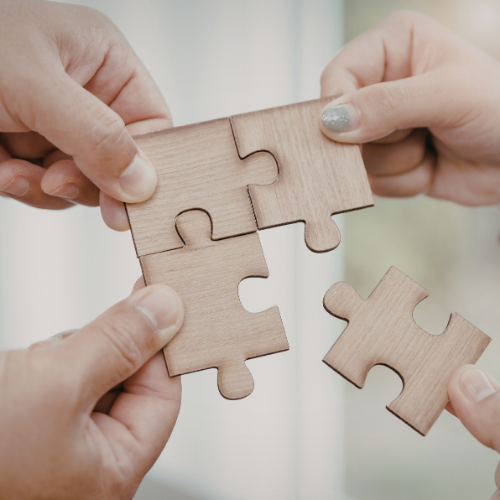Varias manos humanas están ensamblando un rompecabezas de madera, con tres piezas ya unidas y una cuarta a punto de ser colocada.