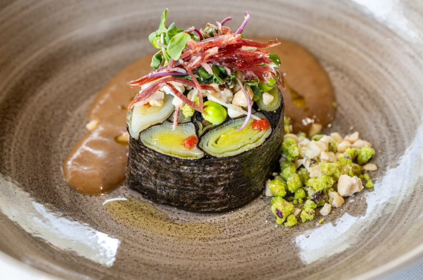 Un plato gourmet presenta un rollo de alga nori relleno de vegetales y coronado con brotes, jamón serrano y frutos secos, acompañado de una salsa marrón y un crumble de pistachos en un plato de cerámica rústica.