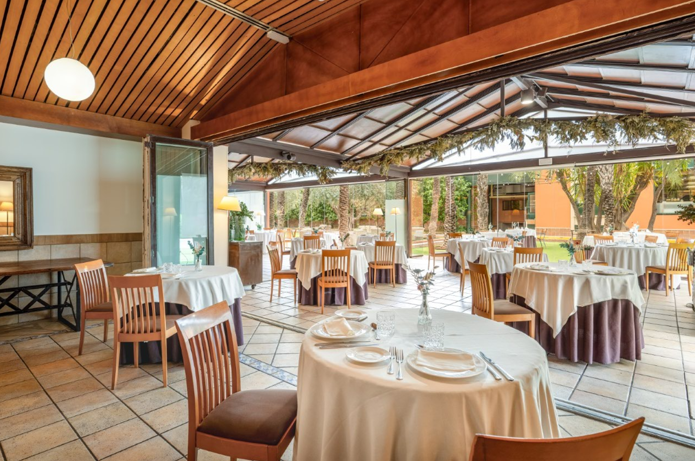 Un elegante comedor de restaurante con mesas puestas y un techo de madera, que se extiende hacia un patio acristalado con vistas a un jardín con palmeras.
