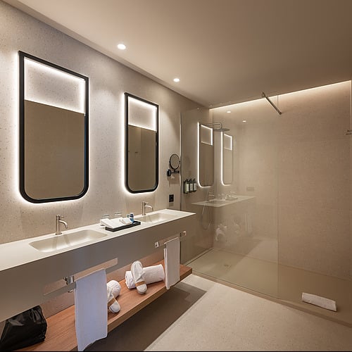 Elegante baño de hotel con doble lavabo, espejo iluminado y ducha de cristal. Ideal para tu Port Hotels: Corporativa.