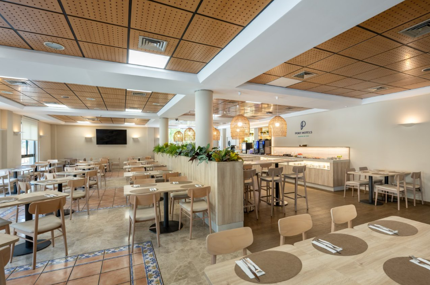 Un moderno y espacioso comedor de hotel con techos de madera perforada, mobiliario claro y una estación de buffet decorada con plantas.