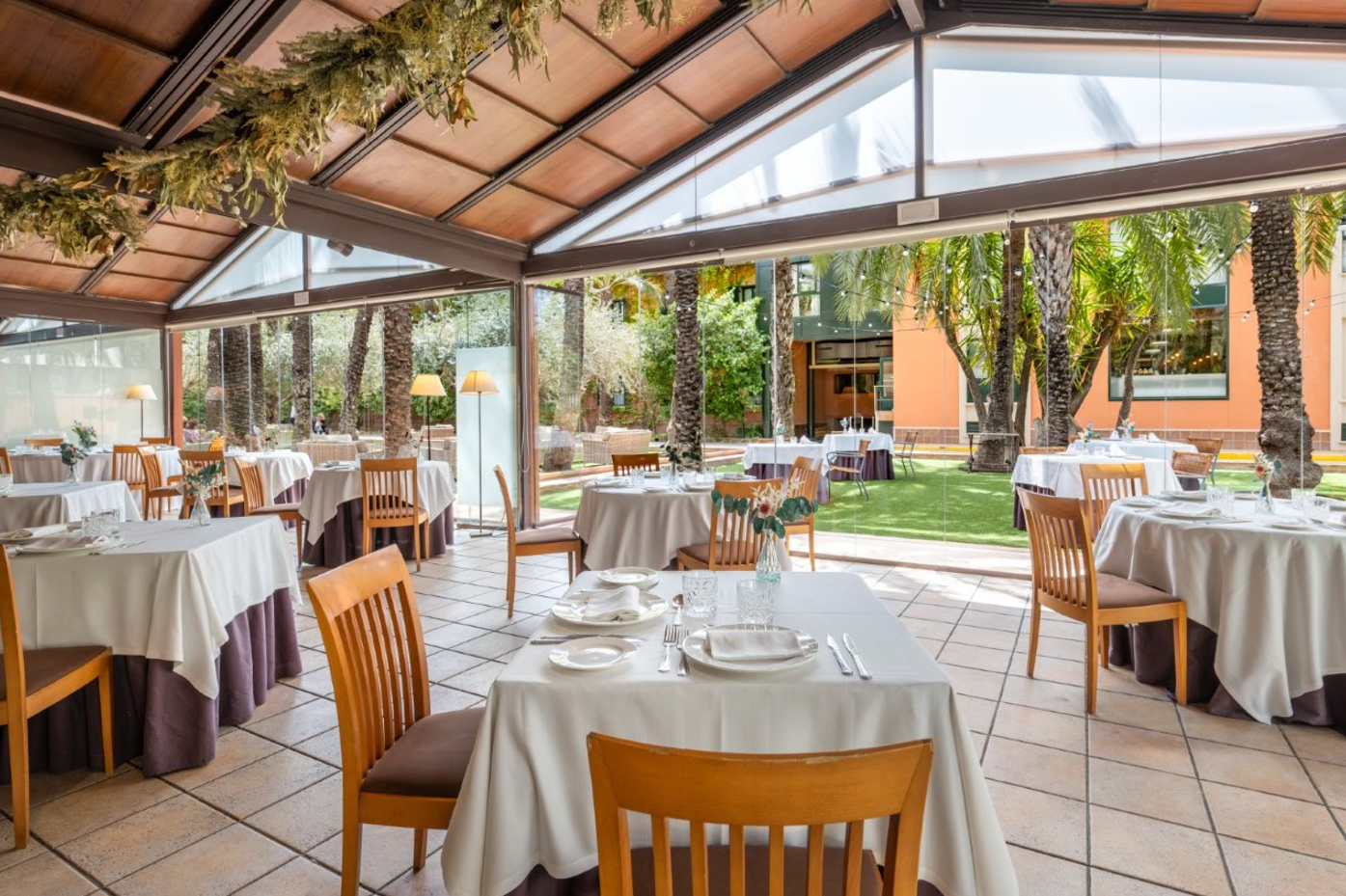 Un luminoso comedor de restaurante con techo y paredes de cristal se abre a un jardín exterior verde con palmeras y mesas.