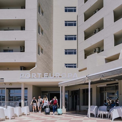 Un grupo de personas con maletas se dirige a la entrada del Hotel Port Europa, una estructura moderna de color claro con balcones y una zona de asientos al aire libre.