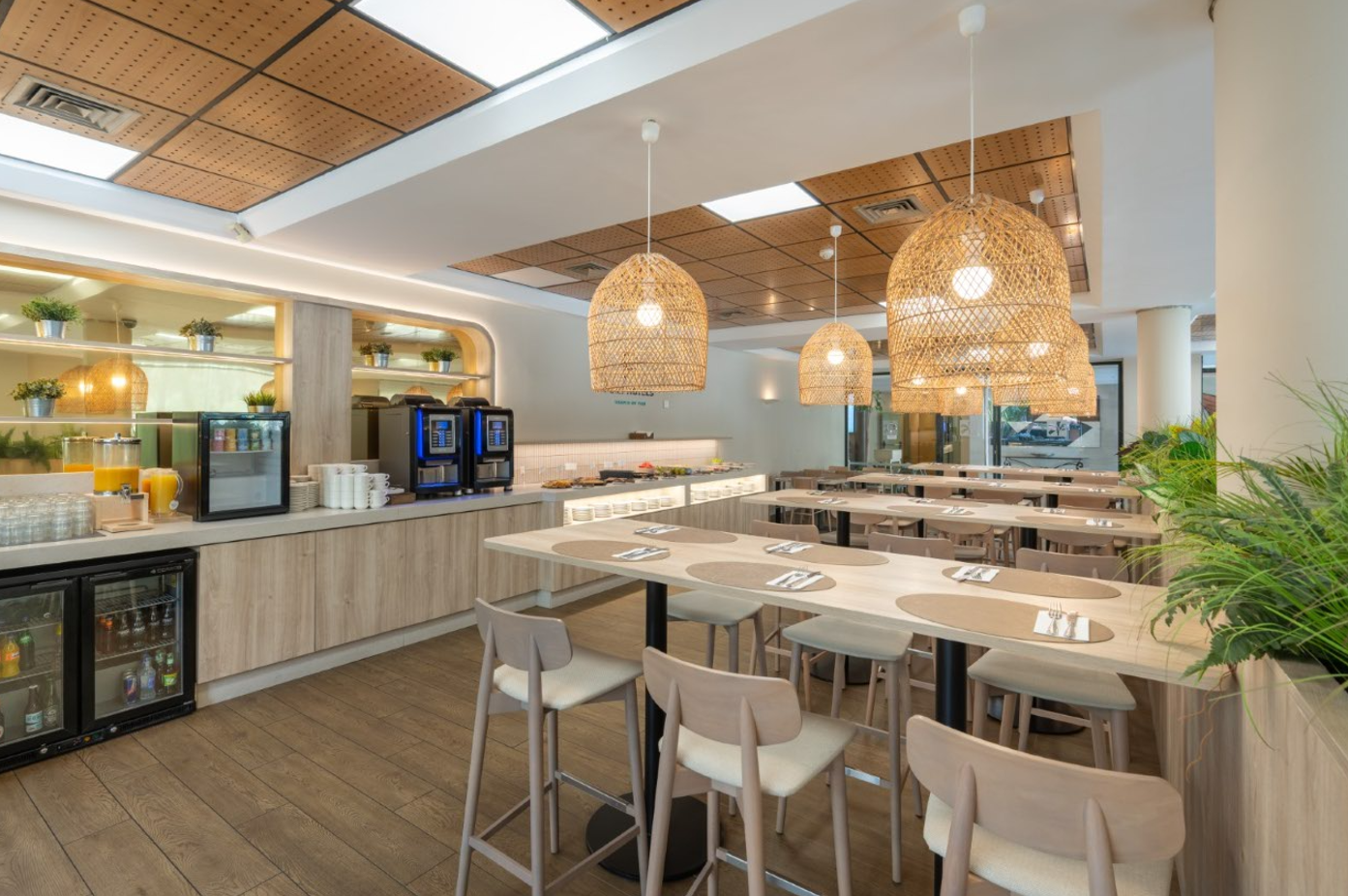 Una moderna y luminosa área de comedor o buffet presenta un mostrador de autoservicio con bebidas y máquinas de café, mesas de madera con sillas altas y lámparas colgantes de mimbre, todo en un ambiente cálido con detalles en madera.
