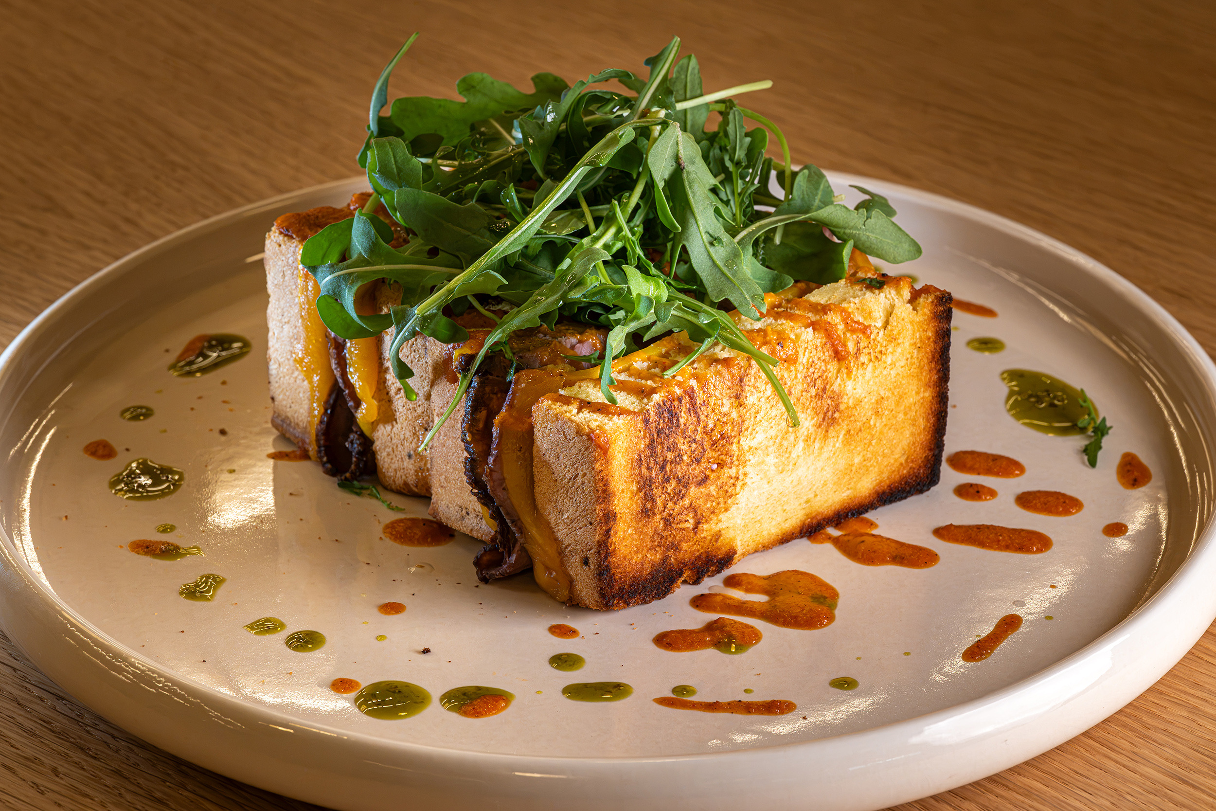 Gourmet sandwich with arugula and melted cheese, served on a plate with sauces. Port Hotels: Corporativa