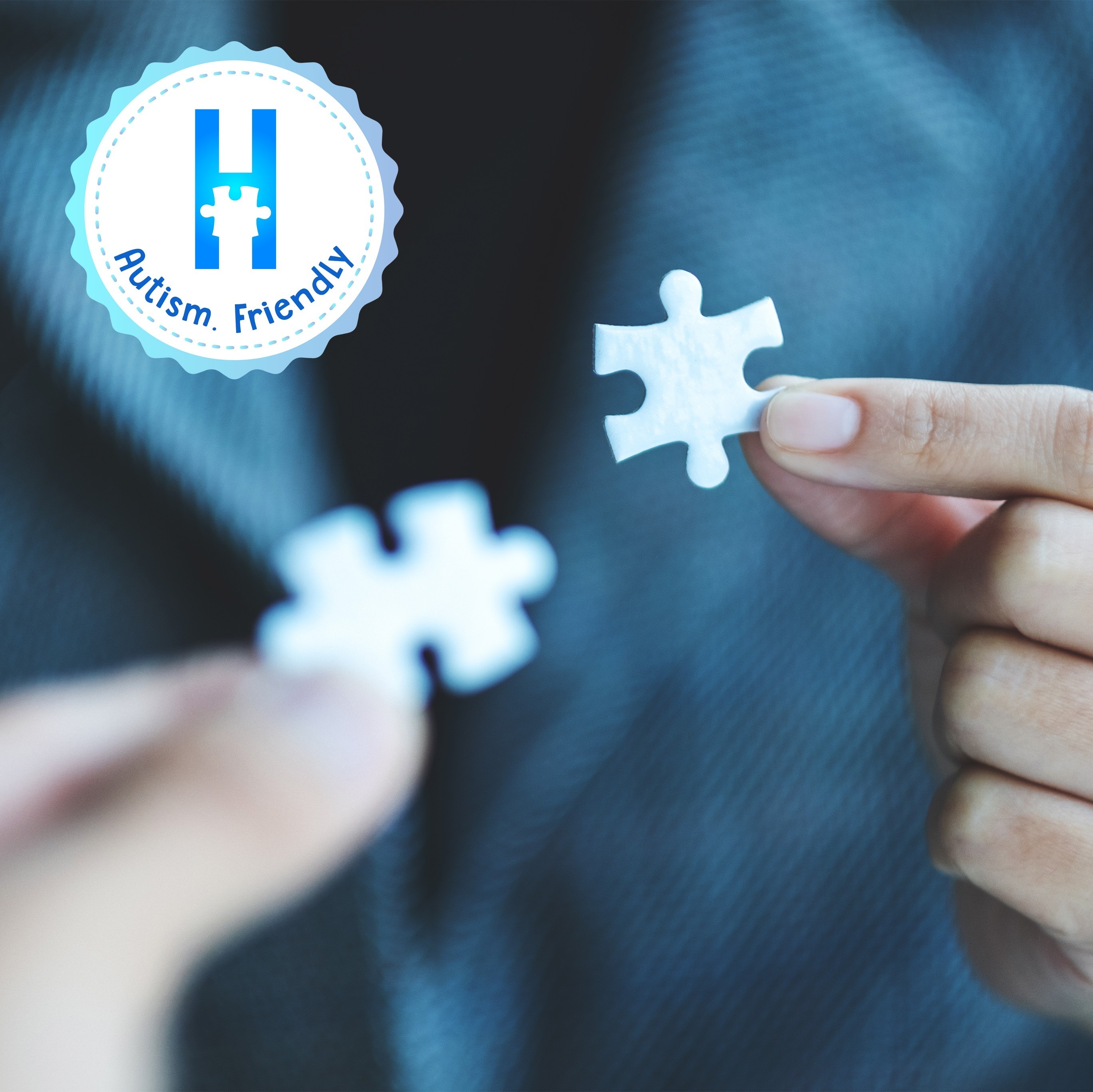 El logotipo presenta una insignia circular azul claro con bordes ondulados, una letra "H" azul con una pieza de rompecabezas blanca en su centro y las palabras "Autism. Friendly." en la parte inferior.
