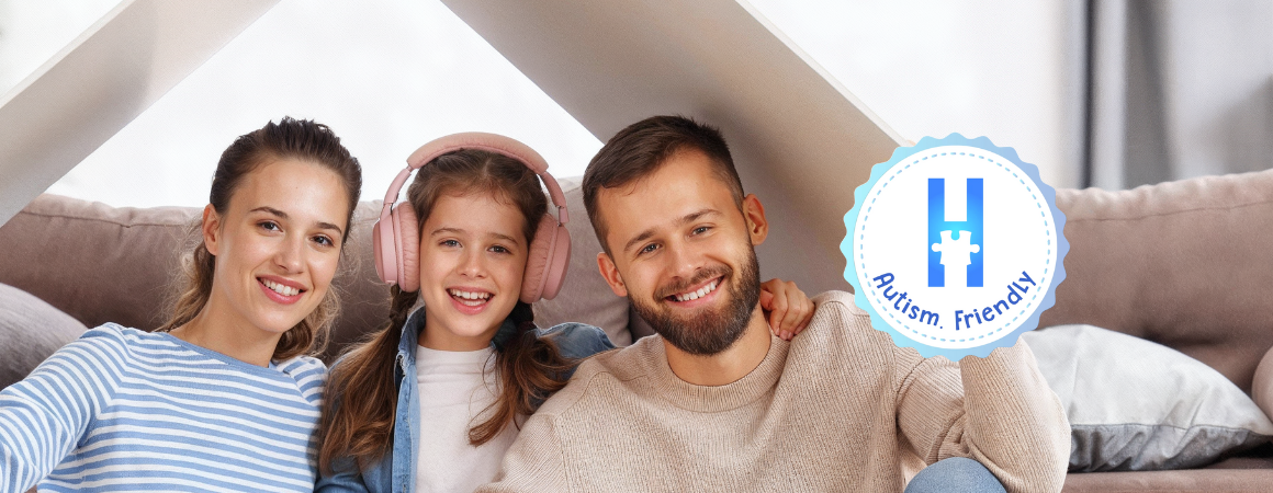 Una familia sonriente, con la hija usando auriculares, aparece junto a un logo de "Autism Friendly" con una 'H' hecha de piezas de rompecabezas.