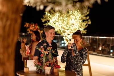 a woman taking a picture of a man sitting at a table with flowers - 