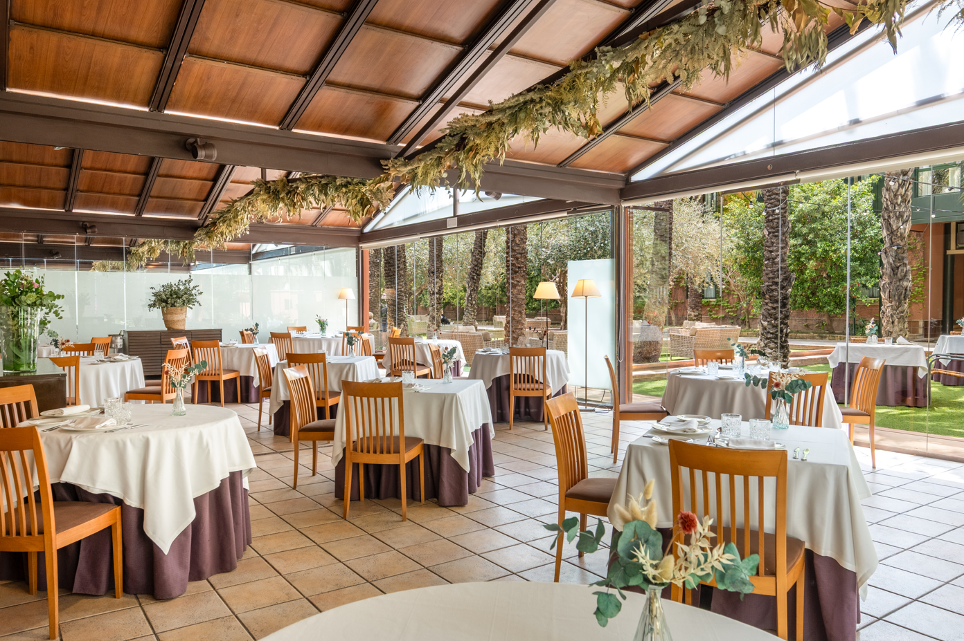 Un elegante comedor con techo de madera y cristal, con mesas dispuestas y vistas a un jardín exuberante.