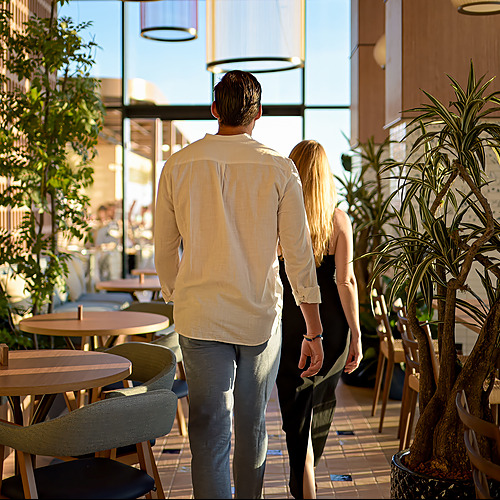 Pareja camina por el restaurante del hotel, con plantas y mesas. Port Hotels: Corporativa