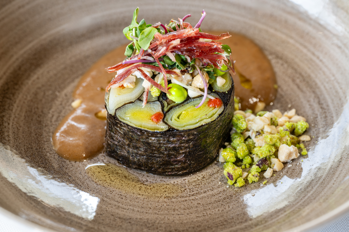 Artfully plated dish: nori-wrapped leeks, prosciutto, microgreens, and savory sauce on a rustic plate.