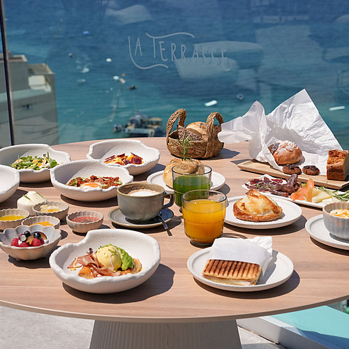 Desayuno gourmet en la terraza 'Port Hotels: Corporativa' con vista al mar y edificios.