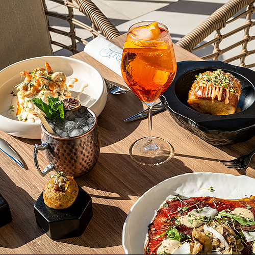 Vista de platos gourmet y cóctel Port Hotels: Corporativa en una terraza soleada con mobiliario de mimbre.