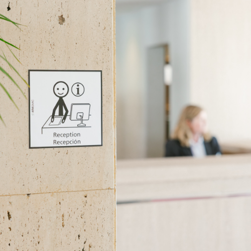 Un letrero de recepción con un pictograma y el símbolo de información (i) está en un pilar de piedra, con una recepcionista desenfocada al fondo.