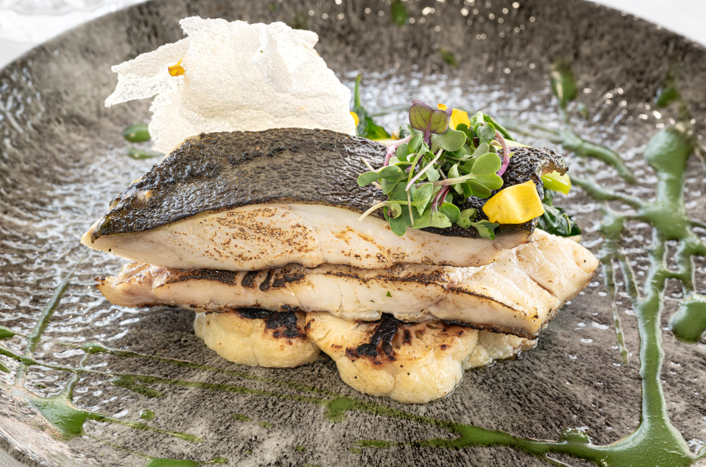 Dos filetes de pescado a la parrilla con piel crujiente se presentan sobre una base de patatas asadas, adornados con microbrotes, un crujiente blanco y salsa verde en un plato texturizado.