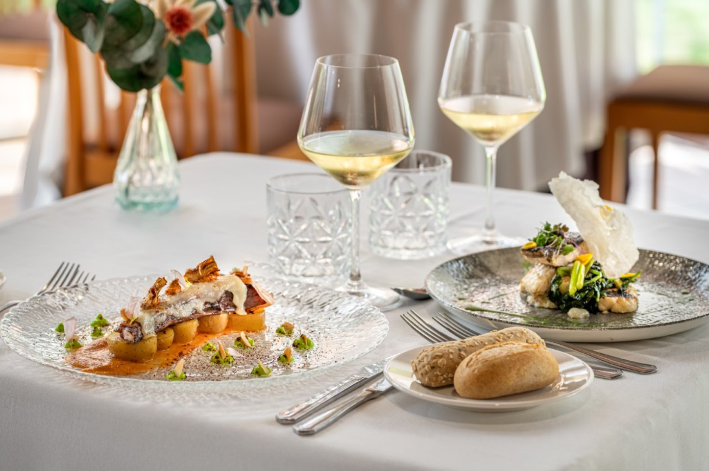 Una mesa de restaurante elegantemente puesta presenta dos platos gourmet, copas de vino blanco y panecillos.