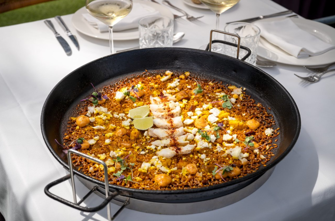 Un gran plato de arroz con mariscos, con trozos de langosta, lima y microbrotes, se presenta en una paellera sobre una mesa de restaurante elegantemente puesta.