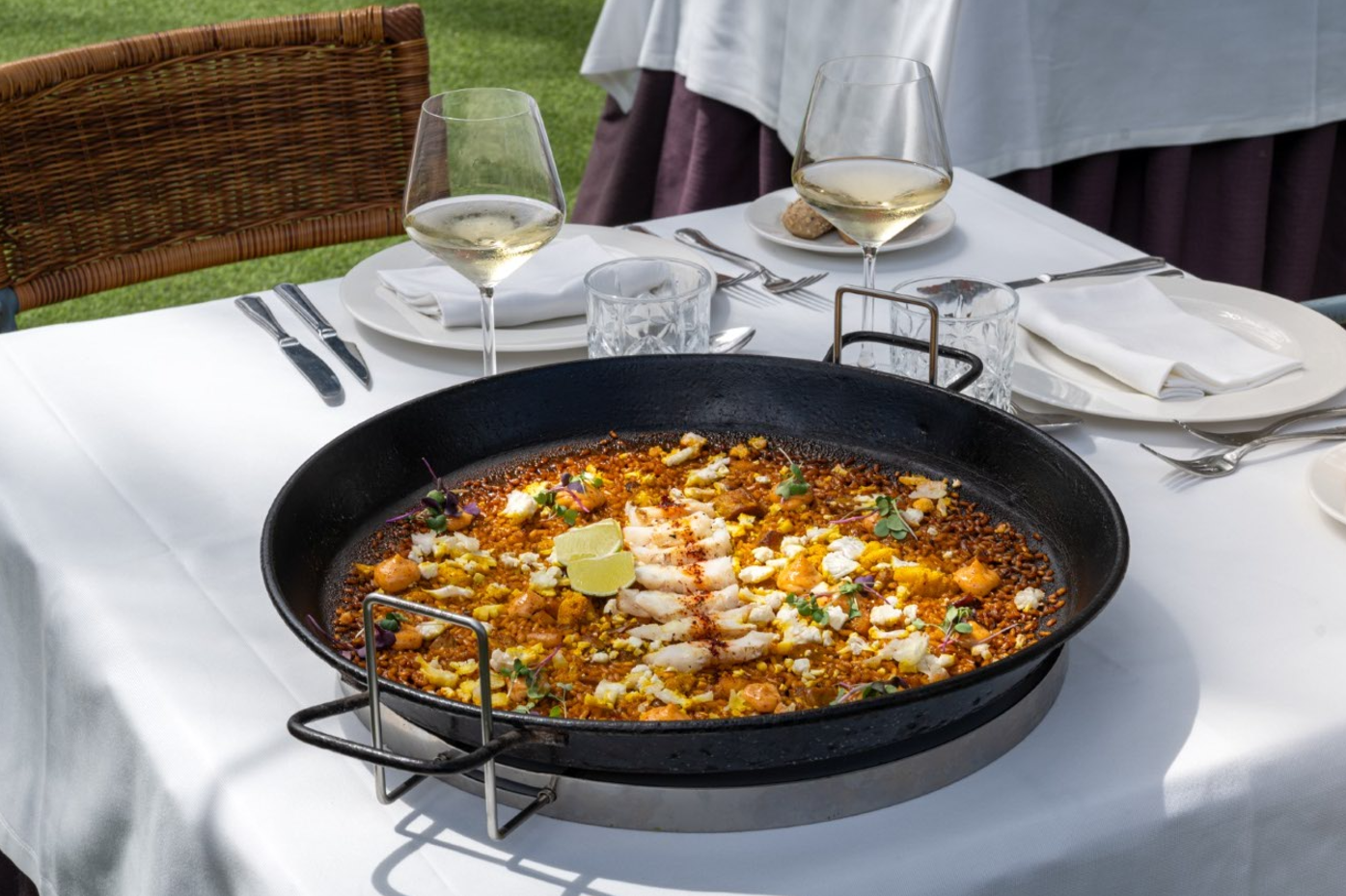 Una paella grande se sirve en una paellera sobre una mesa de restaurante, con dos copas de vino blanco y cubiertos.