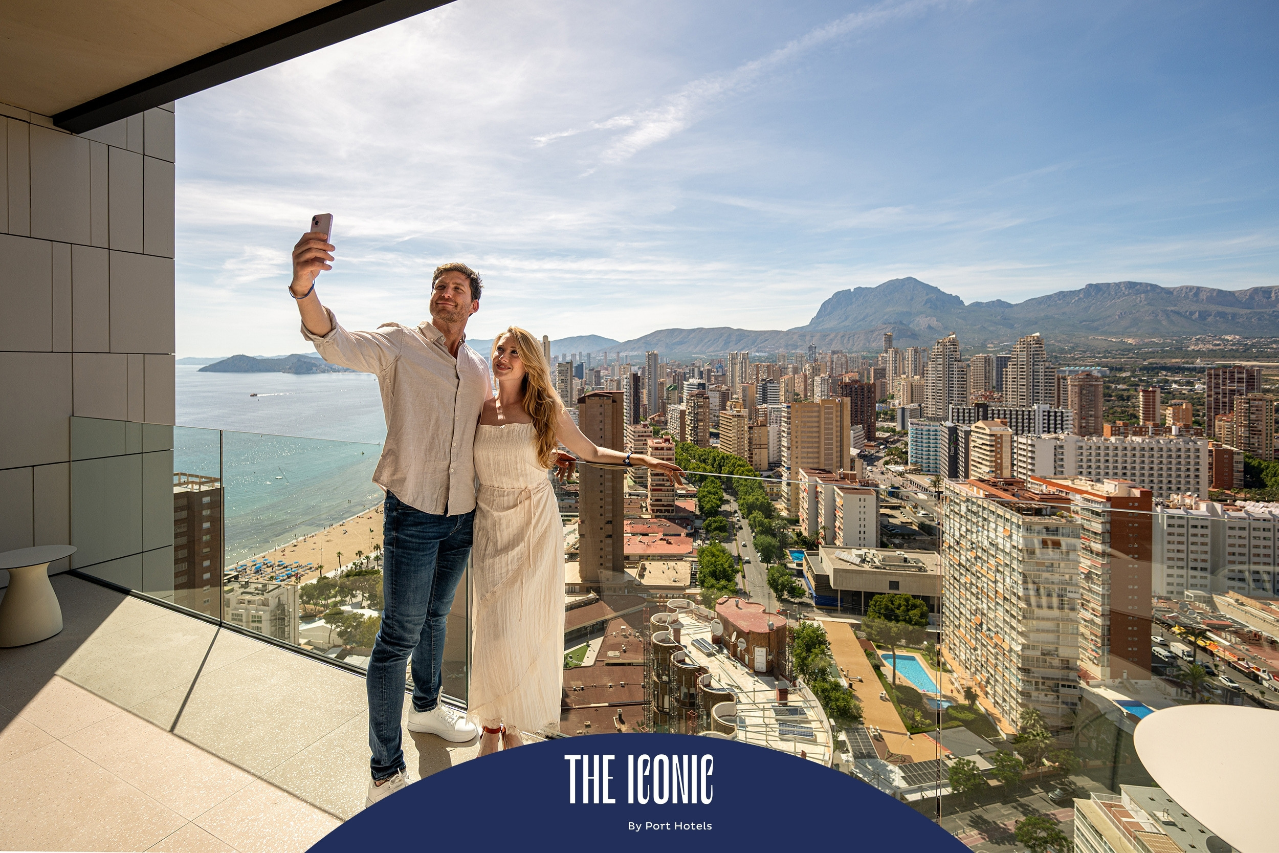 Pareja tomando selfie en balcón con vista a la ciudad y al mar, ideal para Port Hotels: Corporativa.