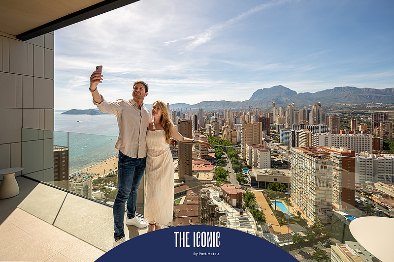 Pareja tomando selfie en balcón con vista a la ciudad y al mar, ideal para Port Hotels: Corporativa.