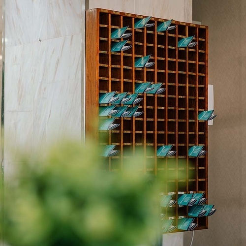 Un tablón de madera con numerosos casilleros, muchos de ellos con tarjetas turquesas, se muestra en un interior, con plantas borrosas en primer plano y una pared de azulejos a la izquierda.