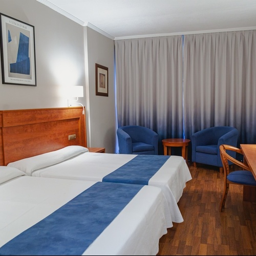 Twin beds in a modern hotel room with blue accent chairs, ideal for Port Hotels: Corporativa stays.