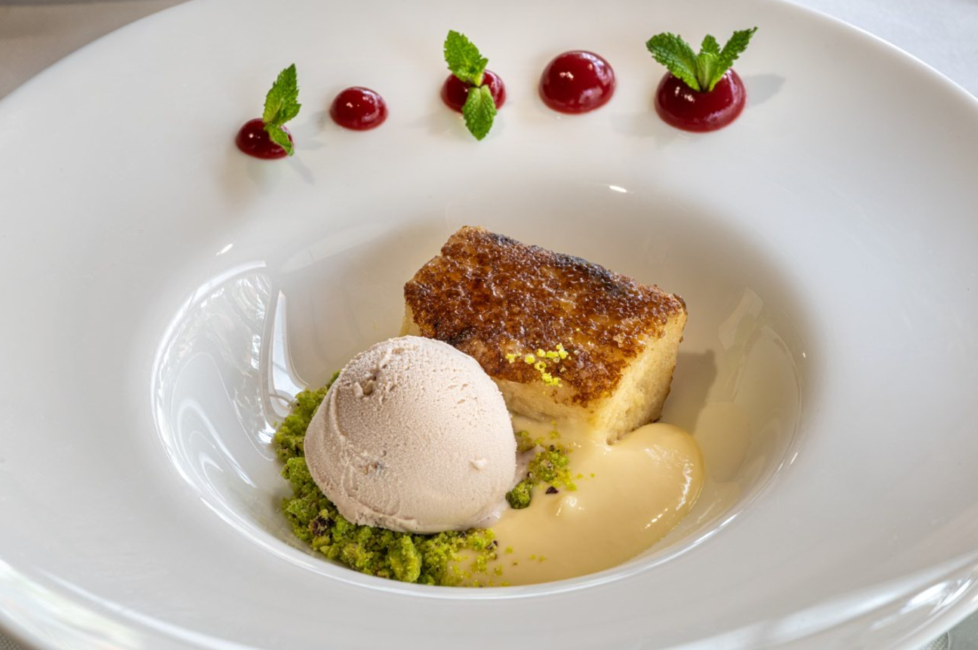 Un elegante postre, que incluye un bizcocho caramelizado, helado, crumble de pistacho y salsa cremosa, está artísticamente presentado en un plato blanco con decoraciones de gelatina roja y hojas de menta.