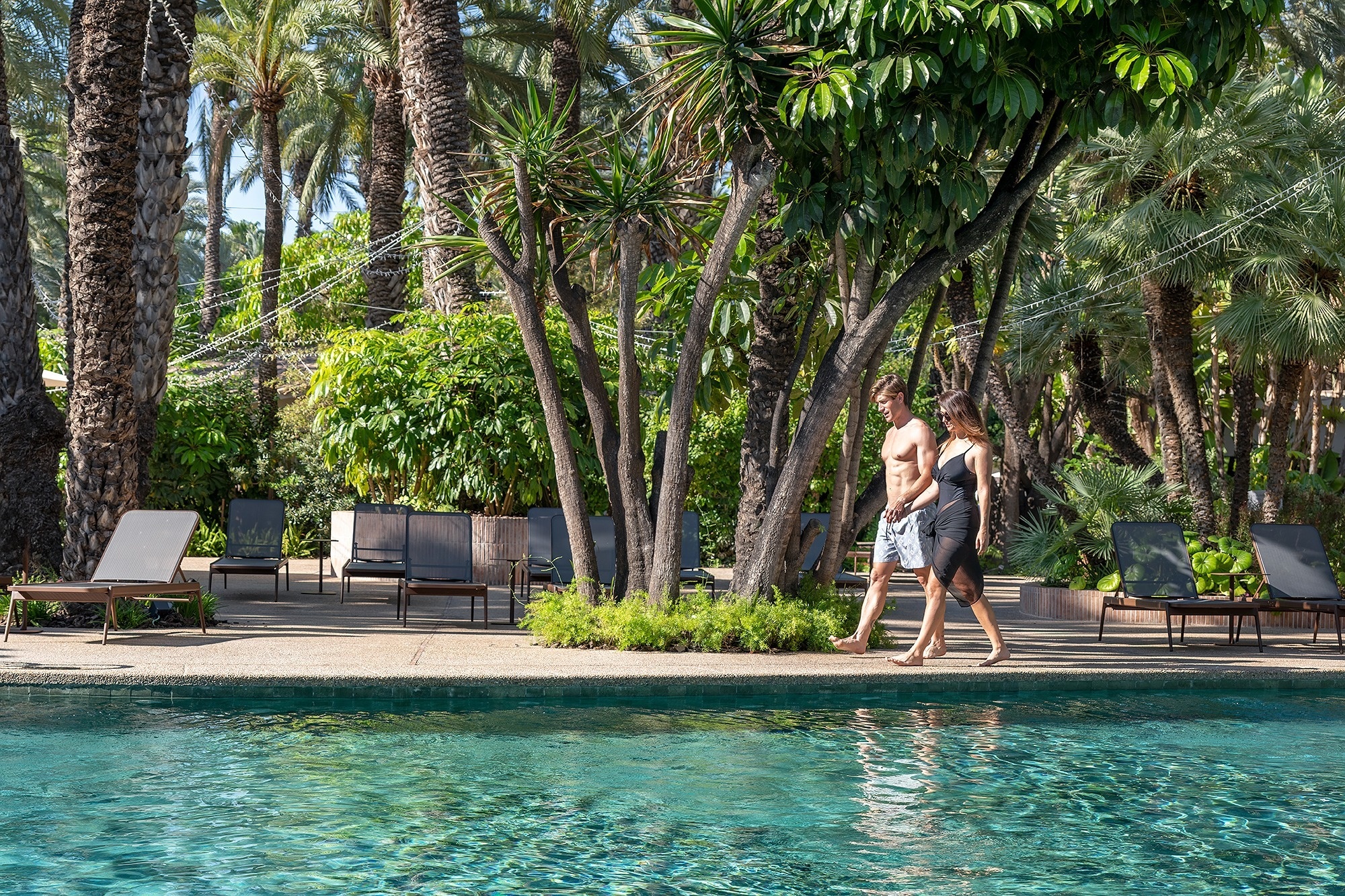 a man and a woman are walking by a pool surrounded by palm trees
